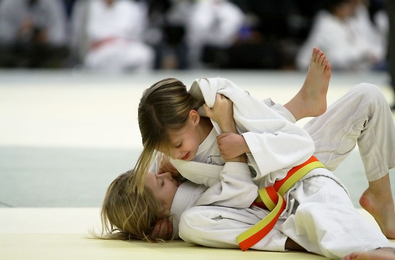 Two girls grapple in judo groundwork, one smiling as she holds on