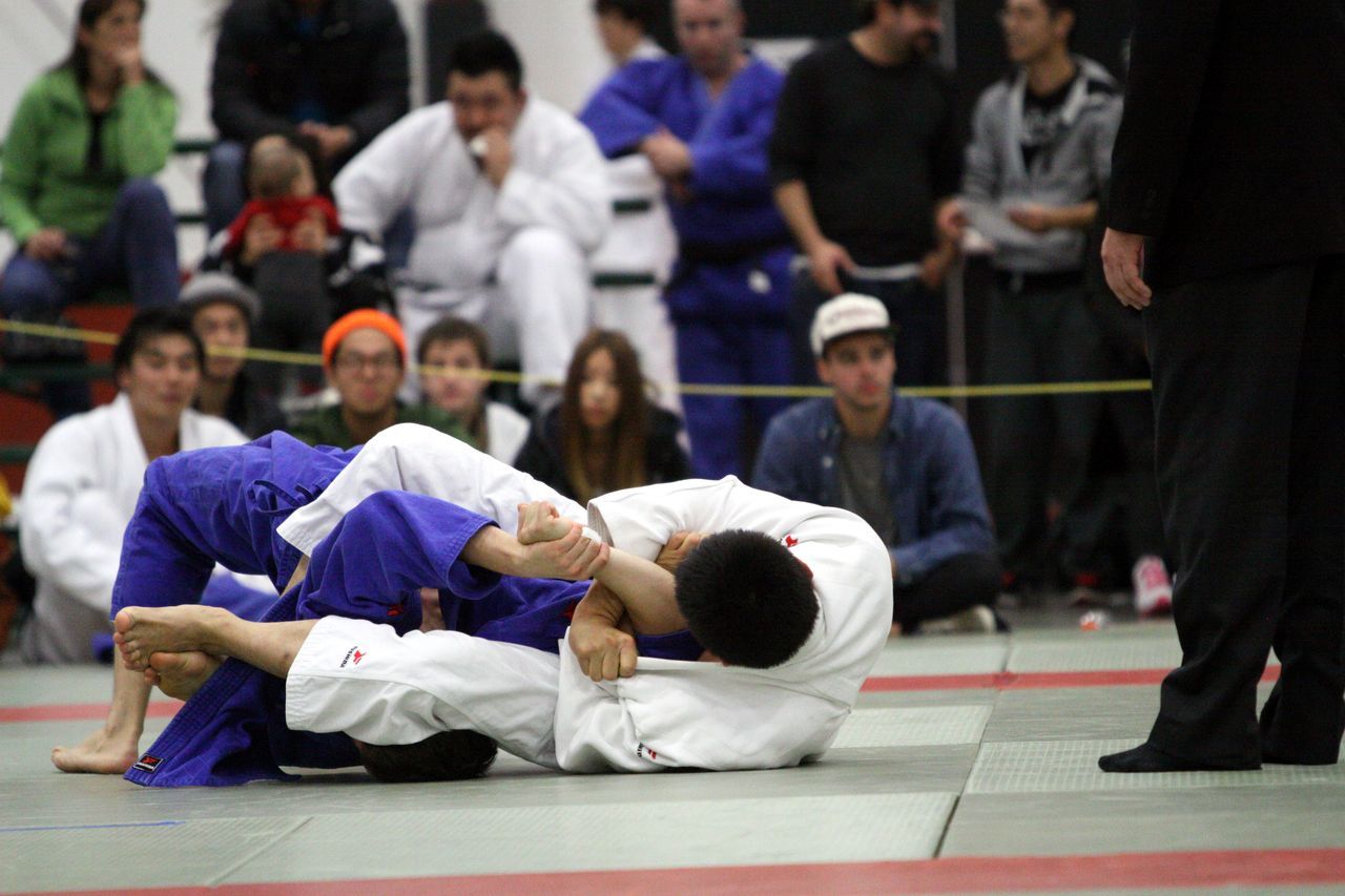 Two judoka work for control in groundwork on the mat as spectators look on