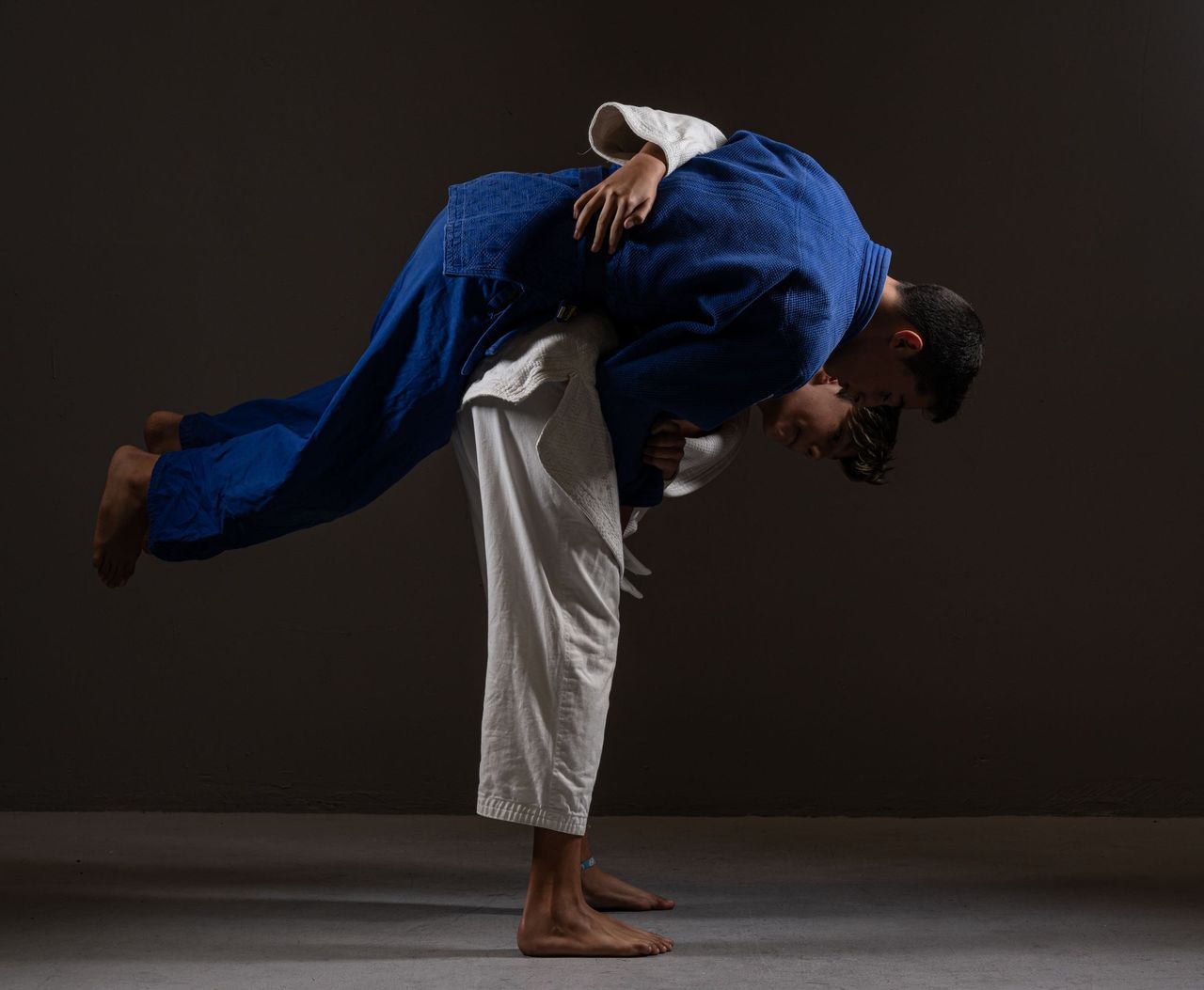 Against a dark wall, a judoka in a white gi lifts his partner in a dark gi off the floor across his back mid-throw
