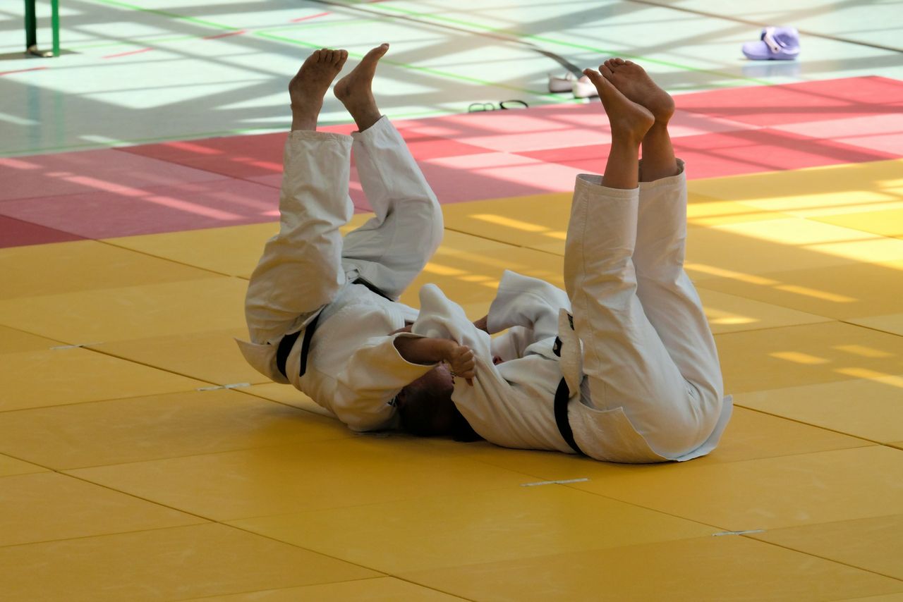 Two judoka in white gis and black belts practise back breakfalls side by side, legs raised straight up